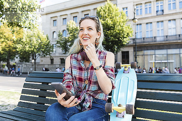 Porträt einer lächelnden jungen Frau auf einer Bank sitzend mit Skateboard und Mobiltelefon