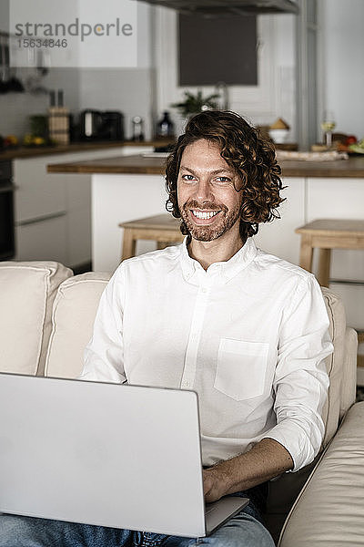Porträt eines lächelnden Mannes mit Laptop auf der Couch zu Hause
