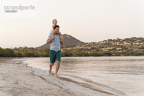 Glücklicher Vater trägt seine Tochter bei Sonnenuntergang am Strand auf den Schultern