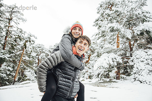 Porträt eines glücklichen jungen Mannes  der seine Freundin im Winterwald Huckepack nimmt