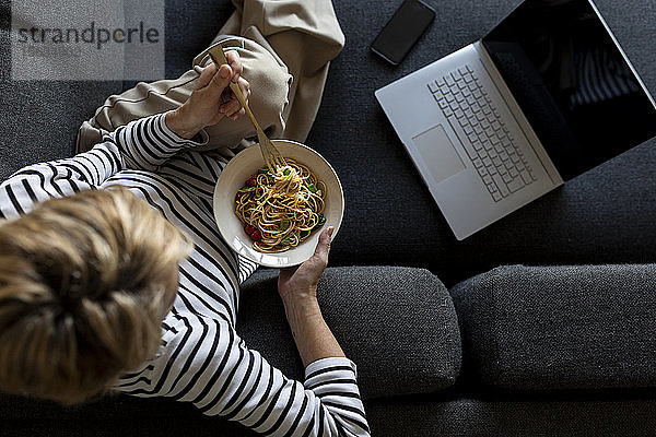 Reife Frau mit Laptop isst zu Hause auf der Couch hausgemachte Nudelgerichte