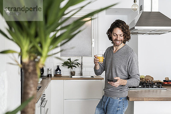 Mann hält zu Hause in der Küche ein Glas Orangensaft und ein Handy