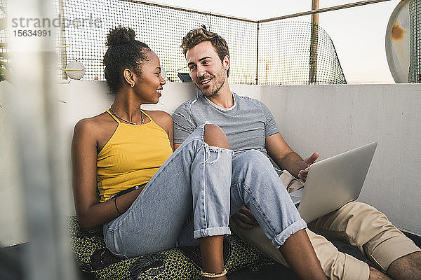 Junges Paar mit Laptop und Kopfhörern sitzt abends auf dem Dach