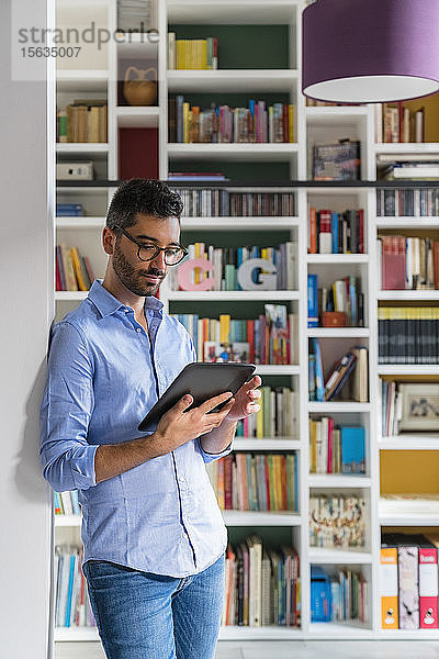 Porträt eines jungen Mannes  der zu Hause mit einem digitalen Tablet vor einem Bücherregal steht