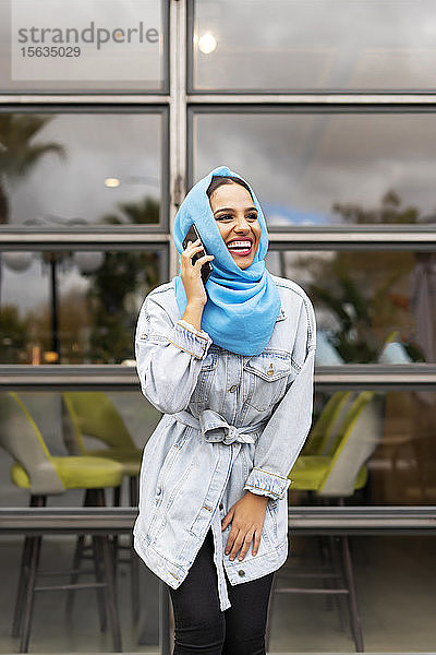 Junge lachende Frau  die einen türkisfarbenen Hijab trägt und vor einem Cafe telefoniert