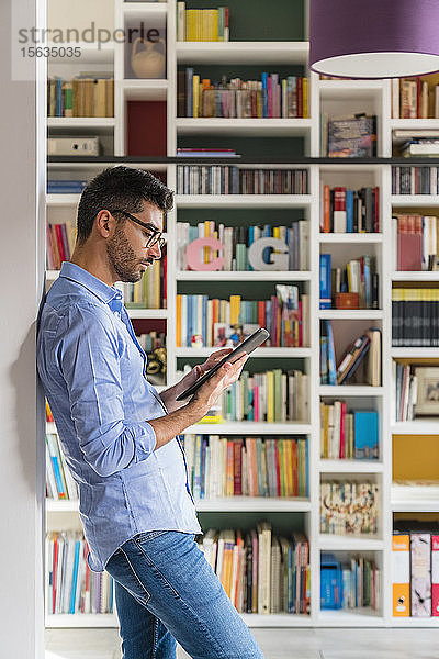 Junger Mann steht zu Hause mit digitalem Tablet vor dem Bücherregal