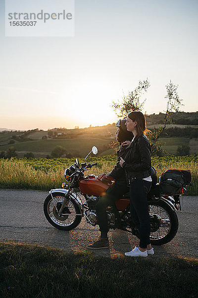 Junges Paar mit einem Oldtimer-Motorrad steht bei Sonnenuntergang auf der Landstraße und schaut auf die Aussicht  Toskana  Italien