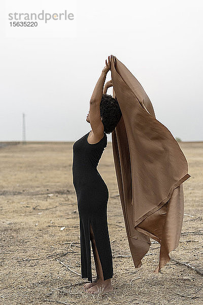Barfüßige junge Frau in schwarzem Kleid steht in trostloser Landschaft und hält eine Decke