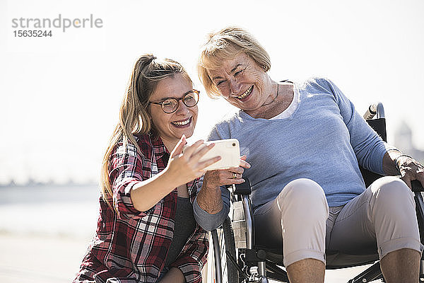 Junge Frau mit ihrer lächelnden Großmutter im Rollstuhl sitzend und mit einem Selfie
