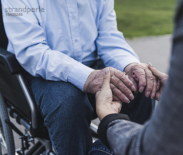 Junge Frau hält Hände eines älteren Mannes  der im Rollstuhl sitzt  Teilansicht