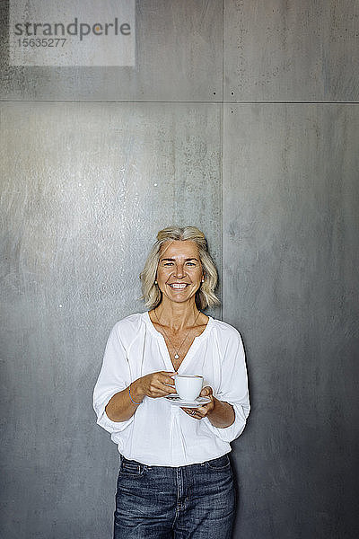 Porträt einer glücklichen  ungezwungenen  reifen Geschäftsfrau  die vor einer grauen Wand steht und eine Kaffeetasse hält
