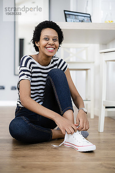 Porträt einer glücklichen jungen Frau  die zu Hause auf dem Boden sitzt und ihre Turnschuhe schnürt