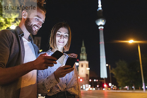 Glückliches Paar benutzt nachts in der Stadt Smartphones  Fernsehturm im Hintergrund  Berlin  Deutschland