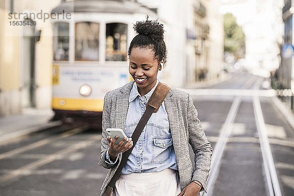Lächelnde junge Frau mit Kopfhörern und Mobiltelefon in der Stadt unterwegs  Lissabon  Portugal
