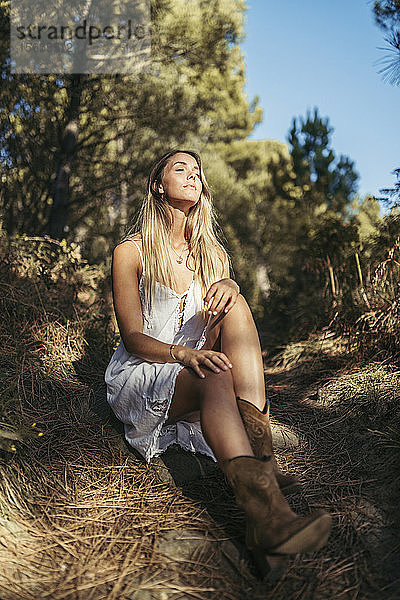 Junge blonde Frau sitzt auf einem Waldweg und genießt das Sonnenlicht mit geschlossenen Augen