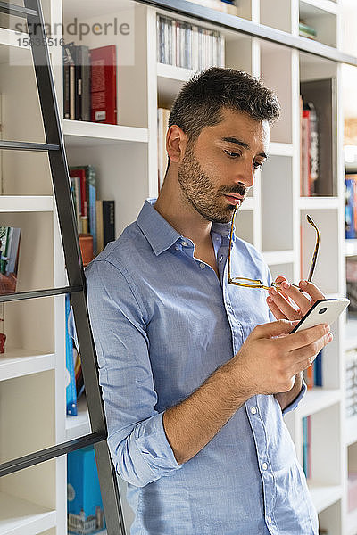 Porträt eines nachdenklichen jungen Mannes  der vor einem Bücherregal steht und auf sein Handy schaut