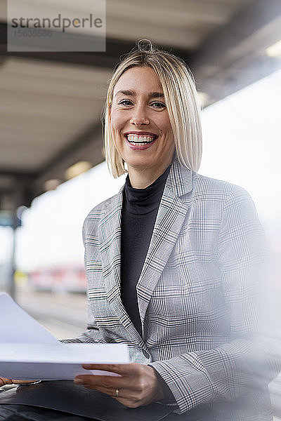 Porträt einer glücklichen jungen Geschäftsfrau mit Papieren am Bahnhof
