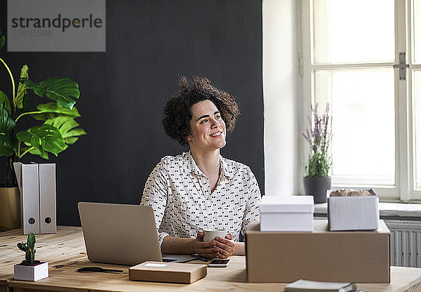 Lächelnde junge Frau sitzt am Schreibtisch mit Kaffeetasse  Laptop und Paketen