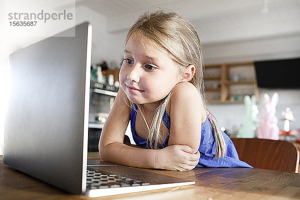 Porträt eines kleinen Mädchens  das zu Hause am Küchentisch lehnt und auf den Laptop schaut