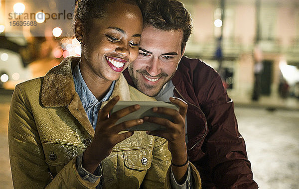 Porträt eines glücklichen jungen Paares  das bei Nacht gemeinsam auf ein Smartphone schaut  Lissabon  Portugal