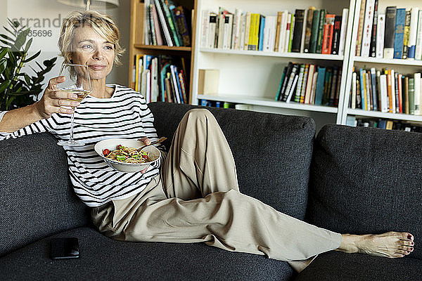 Reife Frau isst zu Hause hausgemachte Nudelgerichte und trinkt Wein auf der Couch