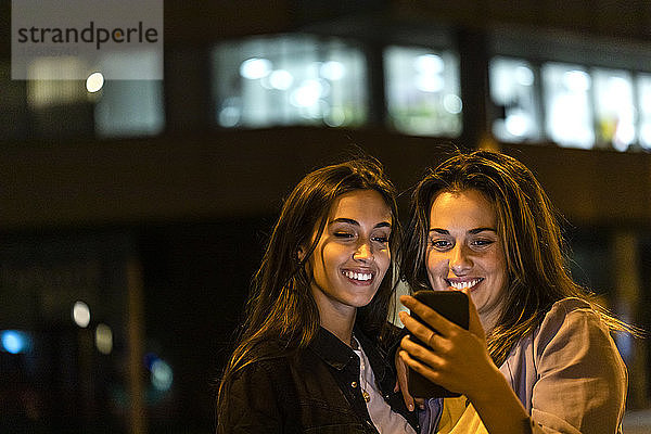 Zwei Freunde benutzen das Smartphone bei Nacht  mit den Lichtern der Stadt im Hintergrund