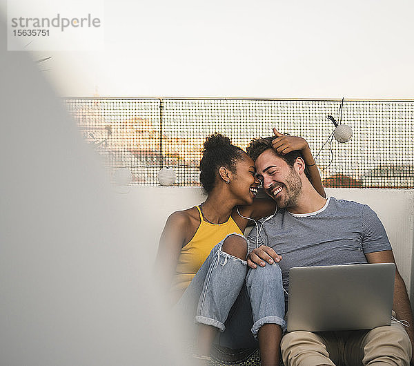 Glückliches junges Paar mit Laptop und Kopfhörern sitzt abends auf dem Dach