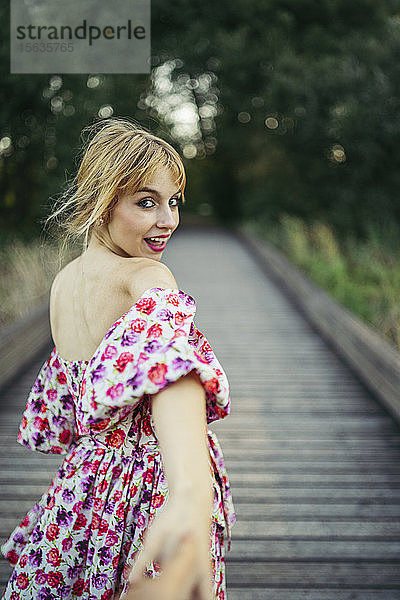 Porträt einer glücklichen jungen Frau in Sommerkleid mit Blumenmuster auf der Promenade  die die Hand hält