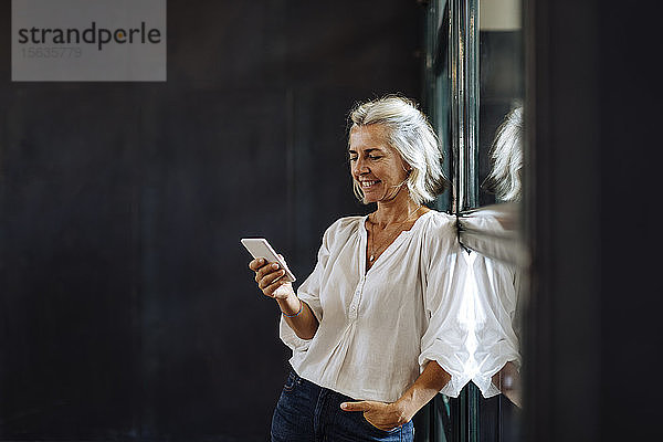 Lächelnde  lässige  reife Geschäftsfrau mit Smartphone am Fenster im Loft-Büro
