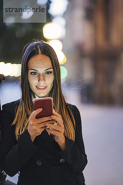 Porträt einer lächelnden jungen Frau  die sich abends an einen Lampenmast lehnt und auf ein Handy schaut