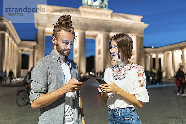 Glückliches Paar benutzt Smartphones am Brandenburger Tor zur blauen Stunde  Berlin  Deutschland