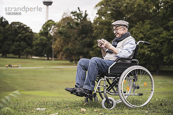Glücklicher älterer Mann sitzt im Rollstuhl auf einer Wiese und benutzt ein Smartphone