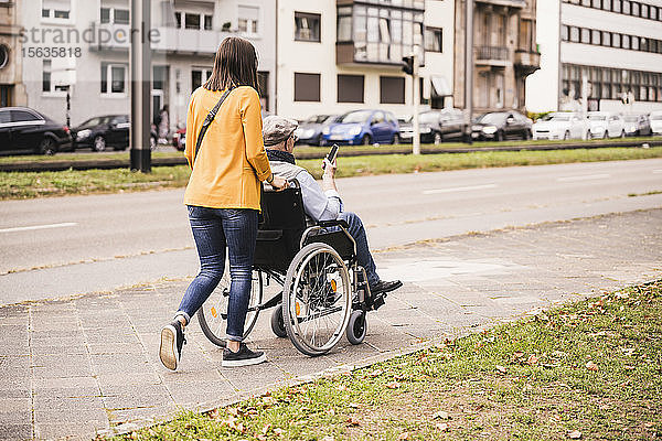 Rückenansicht einer jungen Frau  die einen älteren Mann mit einem Smartphone im Rollstuhl auf dem Bürgersteig schiebt