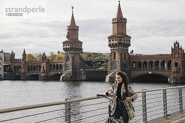 Lächelnde Frau mit Fahrrad und Smartphone in der Stadt an der Oberbaumbrücke  Berlin  Deutschland