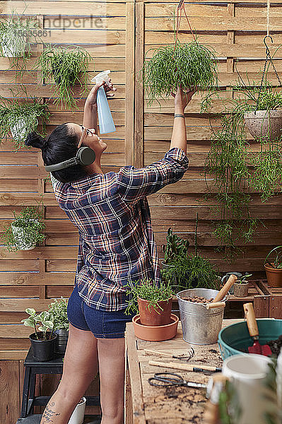 Glückliche Frau  die auf ihrer Terrasse Wasser auf Pflanzen spritzt  während sie Musik hört