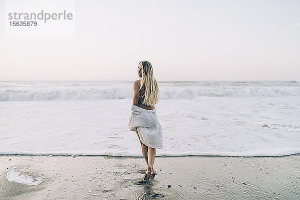 Junge blonde Frau in Bikini und weißem Kleid morgens am Strand