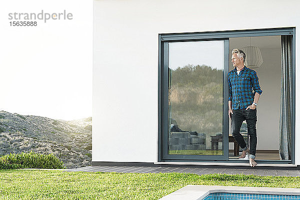 Lässiger Mann  der vor seinem modernen Haus mit Pool steht und auf die Landschaft schaut