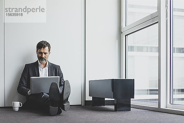 Reifer Geschäftsmann im Büro  der auf dem Boden sitzt und einen Laptop benutzt