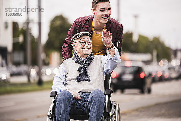 Porträt eines lachenden jungen Mannes  der einen glücklichen älteren Mann im Rollstuhl schiebt