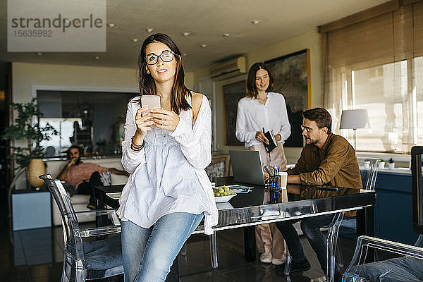 Porträt einer Frau mit Handy und Freunden  die zu Hause im Hintergrund arbeiten