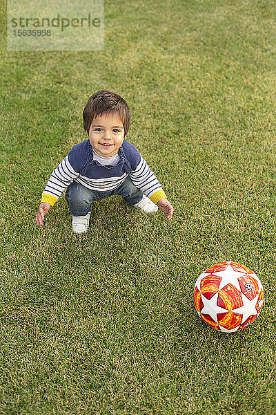 Porträt eines lächelnden kleinen Jungen mit einem Ball  der auf dem Rasen kauert