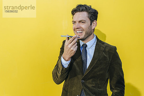 Glücklicher Geschäftsmann mit Smartphone vor gelber Wand