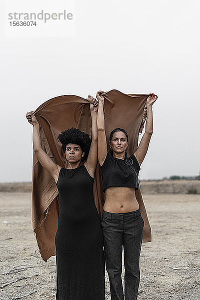 Porträt von zwei schwarz gekleideten Frauen  die zusammen in einer trostlosen Landschaft stehen und sich eine Decke teilen