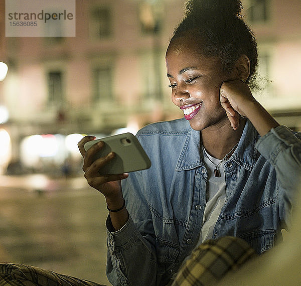 Porträt einer glücklichen jungen Frau  die nachts auf ein Smartphone schaut  Lissabon  Portugal