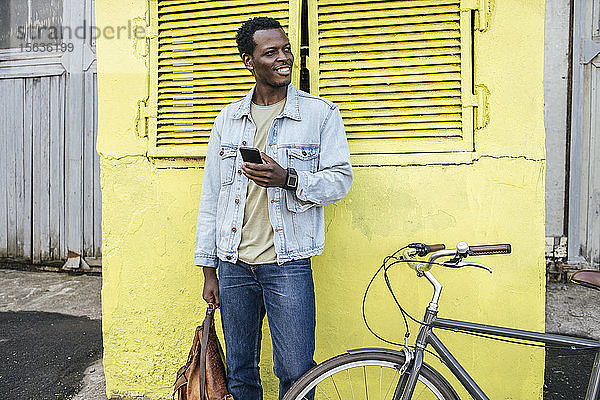Junger Mann mit Smartphone  der vor dem gelben Verteilerkasten wartet