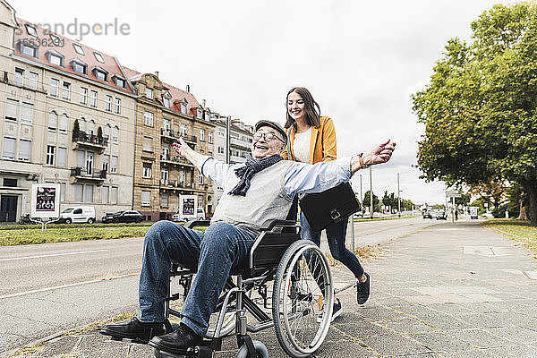 Lächelnde junge Frau schiebt glücklichen älteren Mann im Rollstuhl