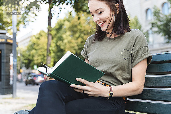 Porträt einer lächelnden jungen Frau  die auf einer Bank sitzt und ein Buch liest