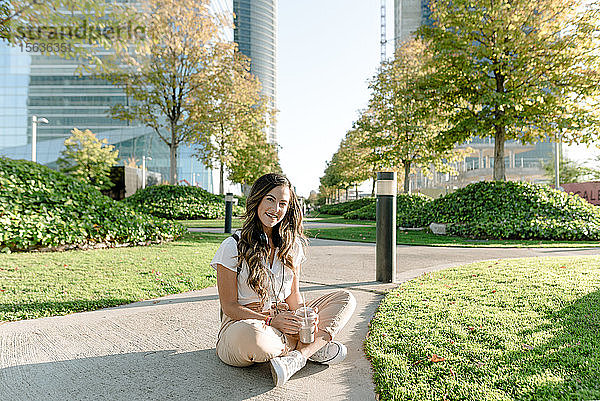 Junge Frau sitzt im Park  trinkt Milchshake und schaut in die Kamera
