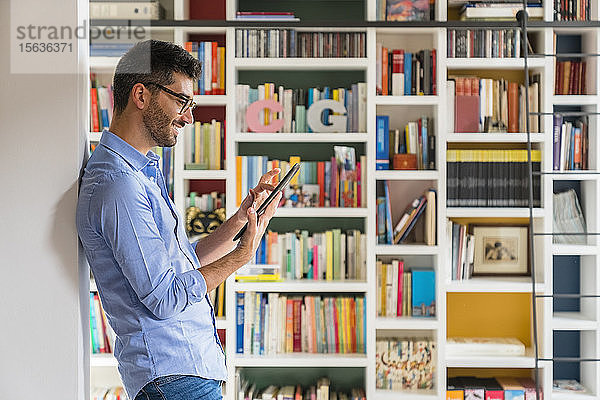 Lächelnder junger Mann steht zu Hause mit digitalem Tablet vor dem Bücherregal