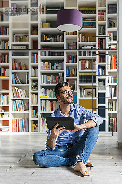 Porträt eines barfüssigen jungen Mannes  der mit einem digitalen Tablett vor Bücherregalen auf dem Boden sitzt